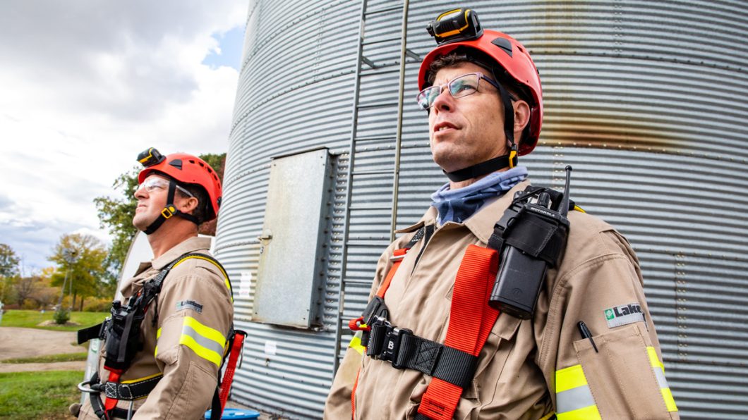 Two men in safety gear standing in front of a grain bin