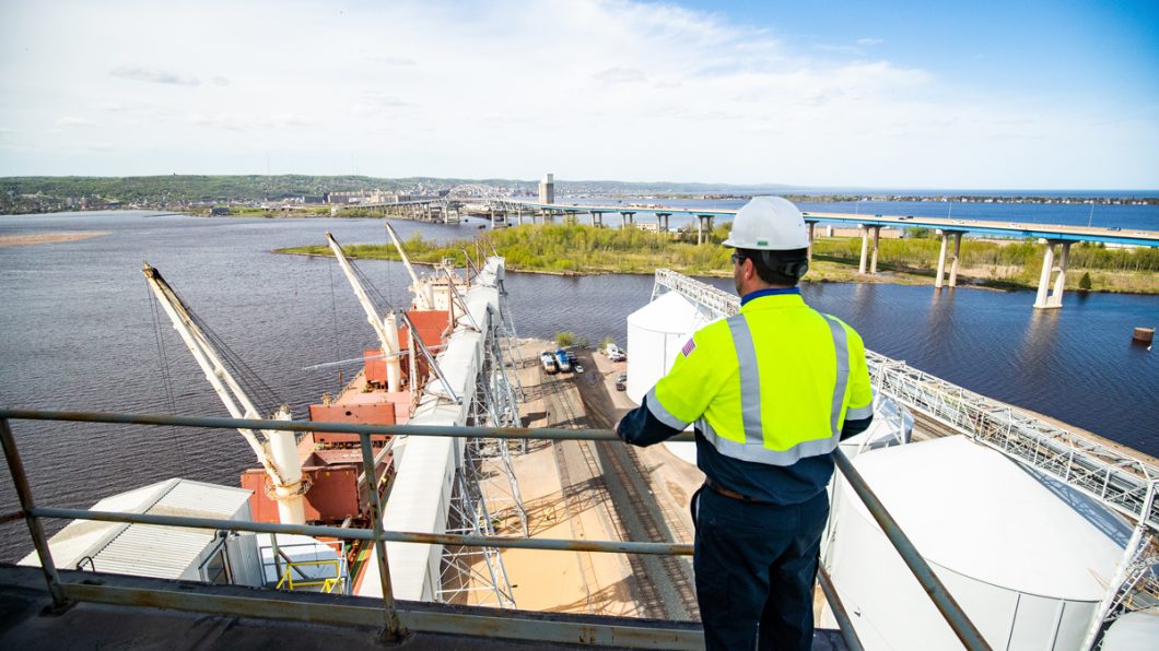 Man in a hard hat and reflective clothes standing on a high platform overlooking a port